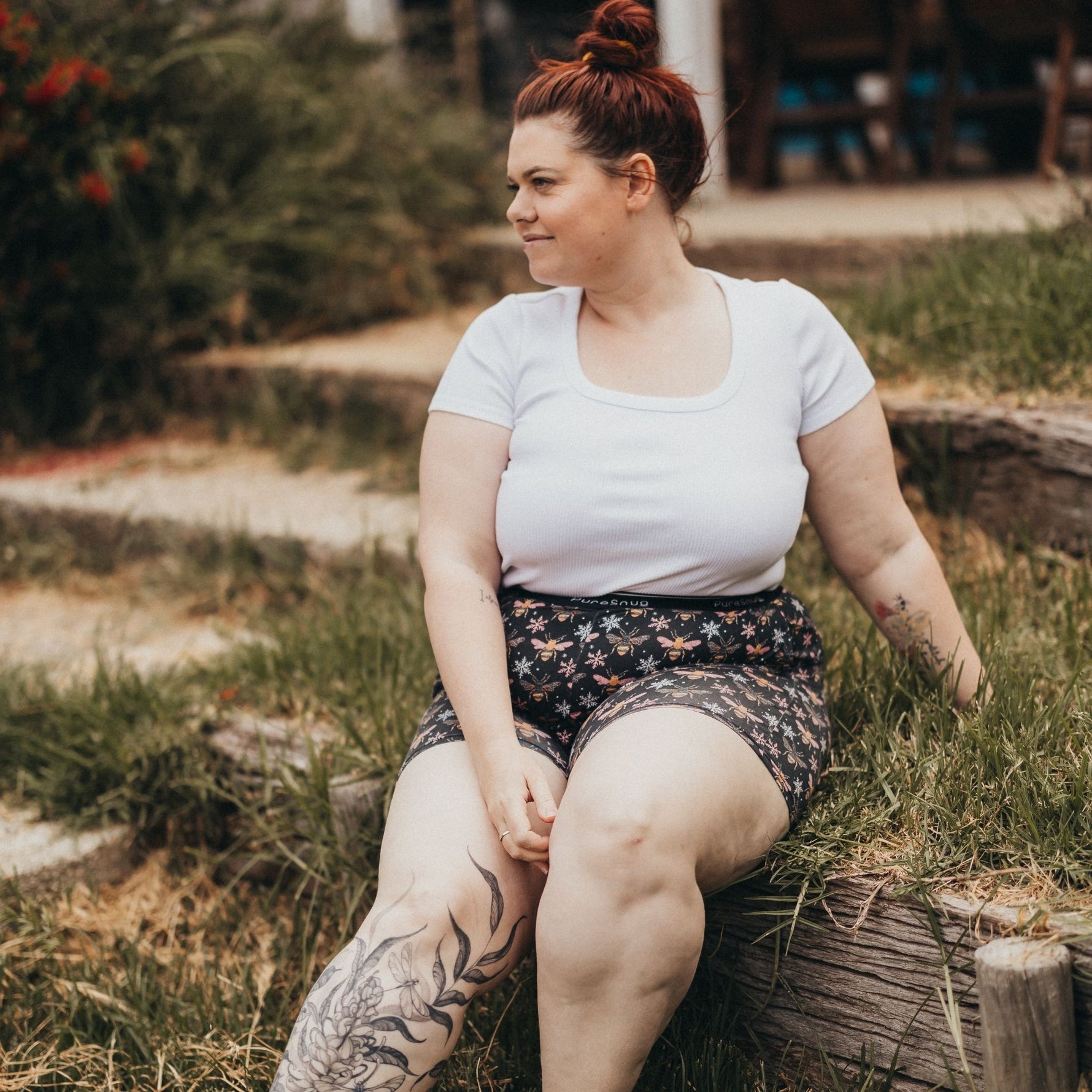 A woman sits on a grassy area beside wooden logs, wearing a white top and bees shorts.