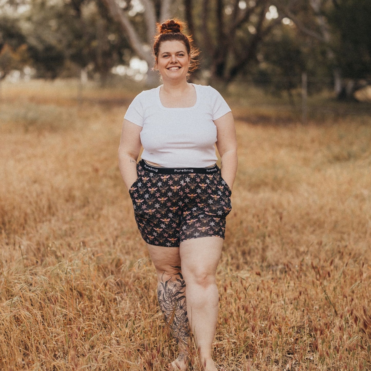 A person wearing a white shirt and patterned lounge shorts walks confidently through a sunlit grass field, surrounded by trees.