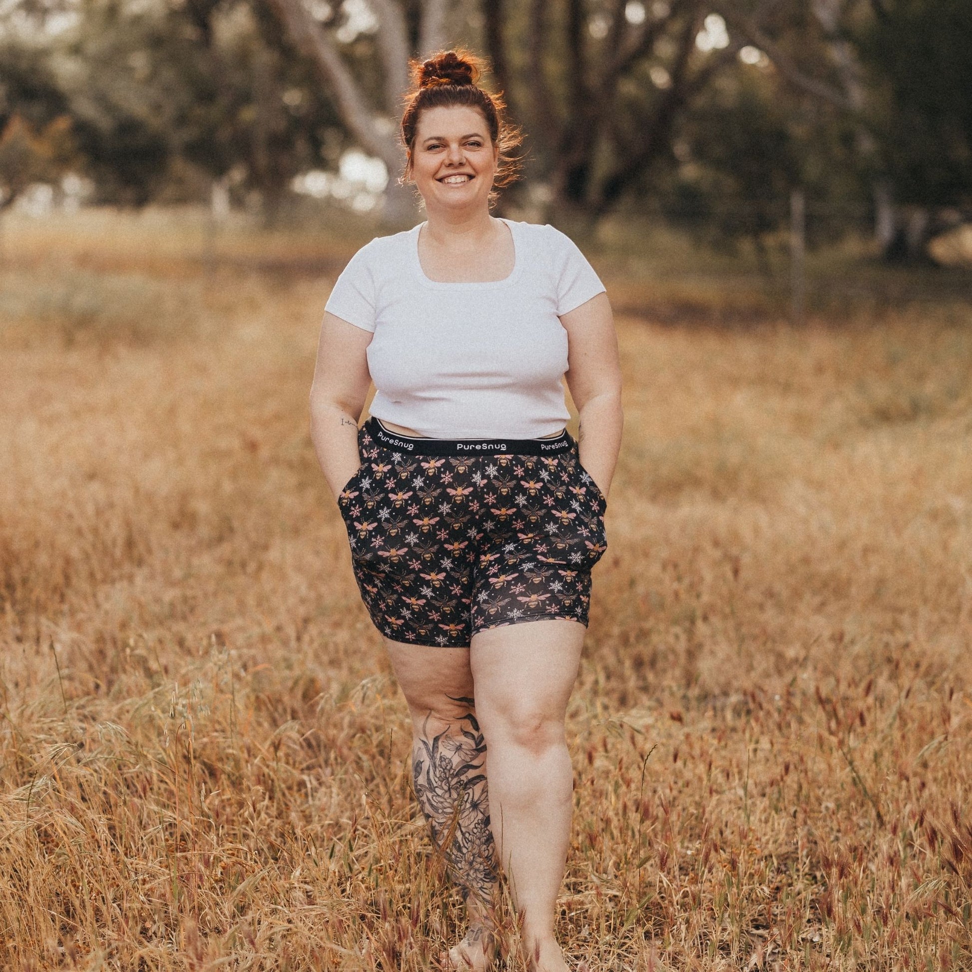 A person wearing a white shirt and patterned lounge shorts walks confidently through a sunlit grass field, surrounded by trees.