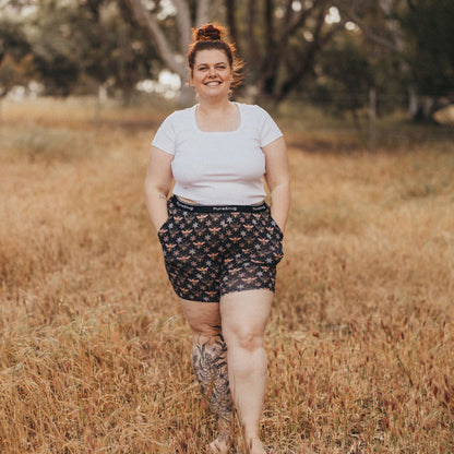 A person wearing a white shirt and patterned lounge shorts walks confidently through a sunlit grass field, surrounded by trees.
