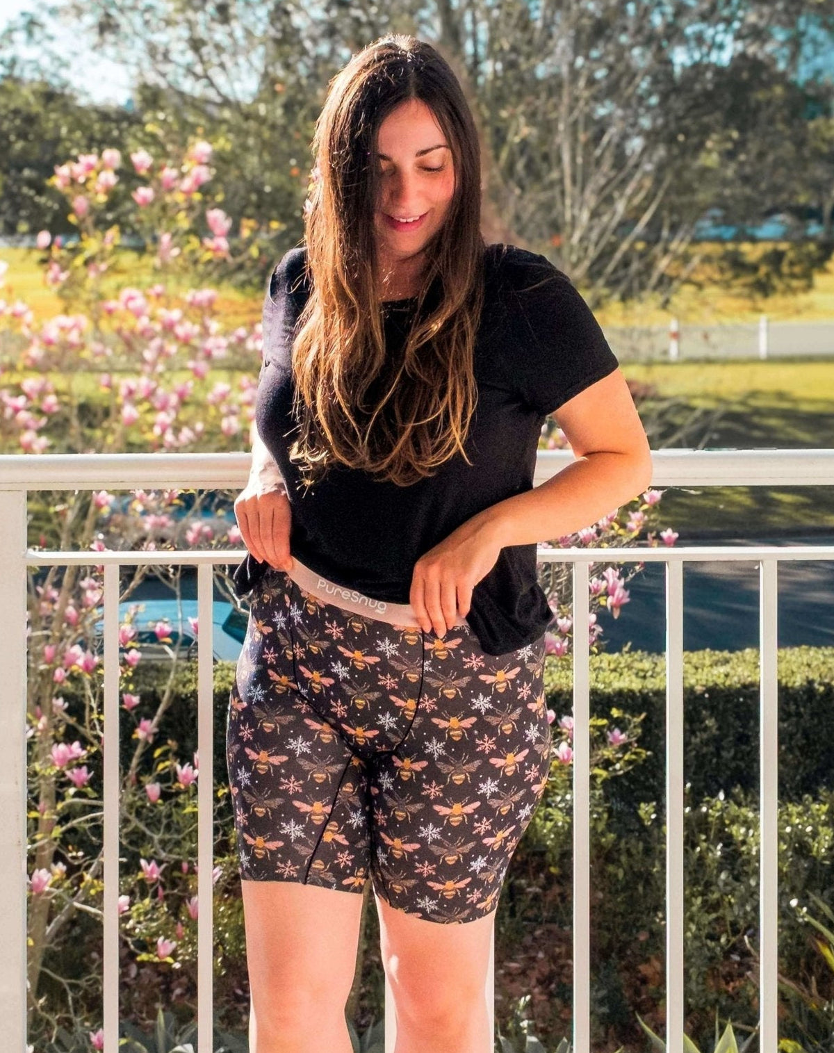 A woman stands on a balcony wearing a black t-shirt and Pure Snug biker briefs in Bees print. The background shows a sunny outdoor park with green grass, trees, and pink flowers.