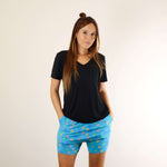 A woman wearing a black PureSnug  v-neck shirt and puresnug lounge shorts in aqua colour with paw print, standing with hands in pockets.