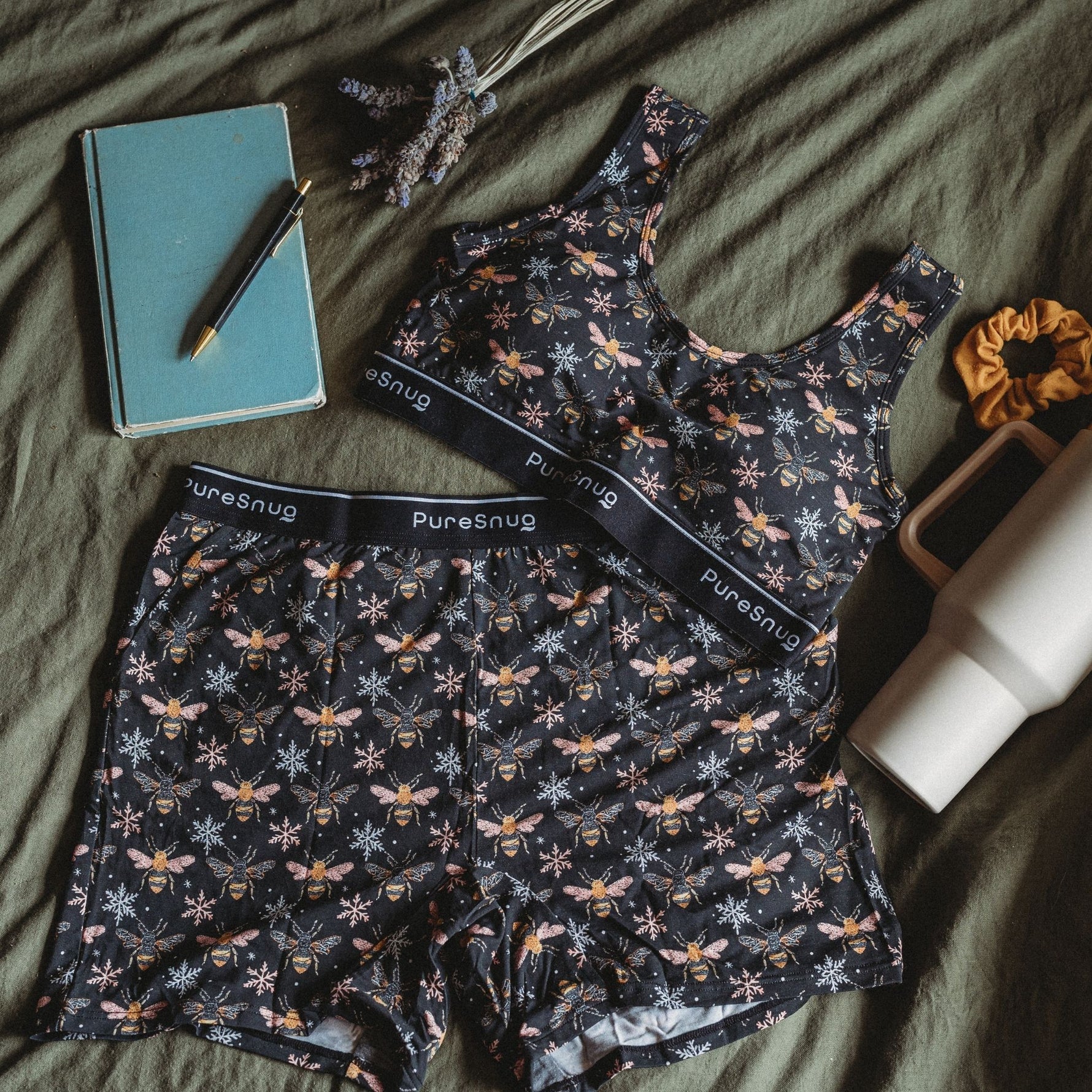 A lounge wear set featuring a crop top bra and lounge shorts lies on an olive green bedspread, accompanied by a notebook, pen, and water bottle.