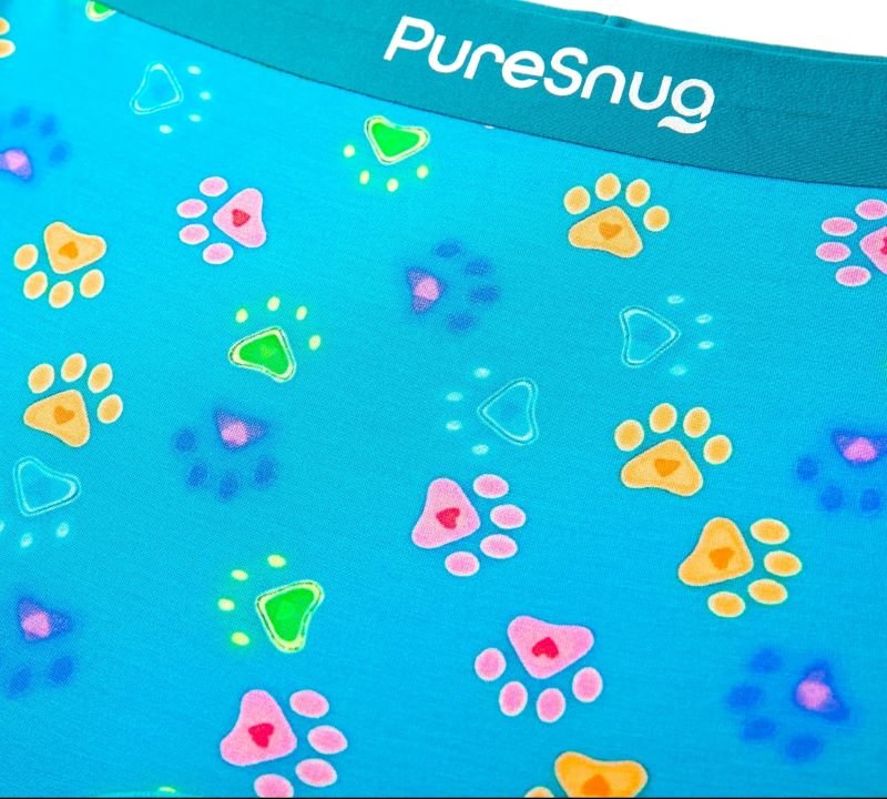 Close-up of bright blue fabric featuring colorful paw prints and geometric shapes with the "PureSnug" logo at the top.