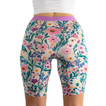 A model in a white top showcases colorful, floral-patterned bike shorts, highlighting vibrant blooms and greenery.