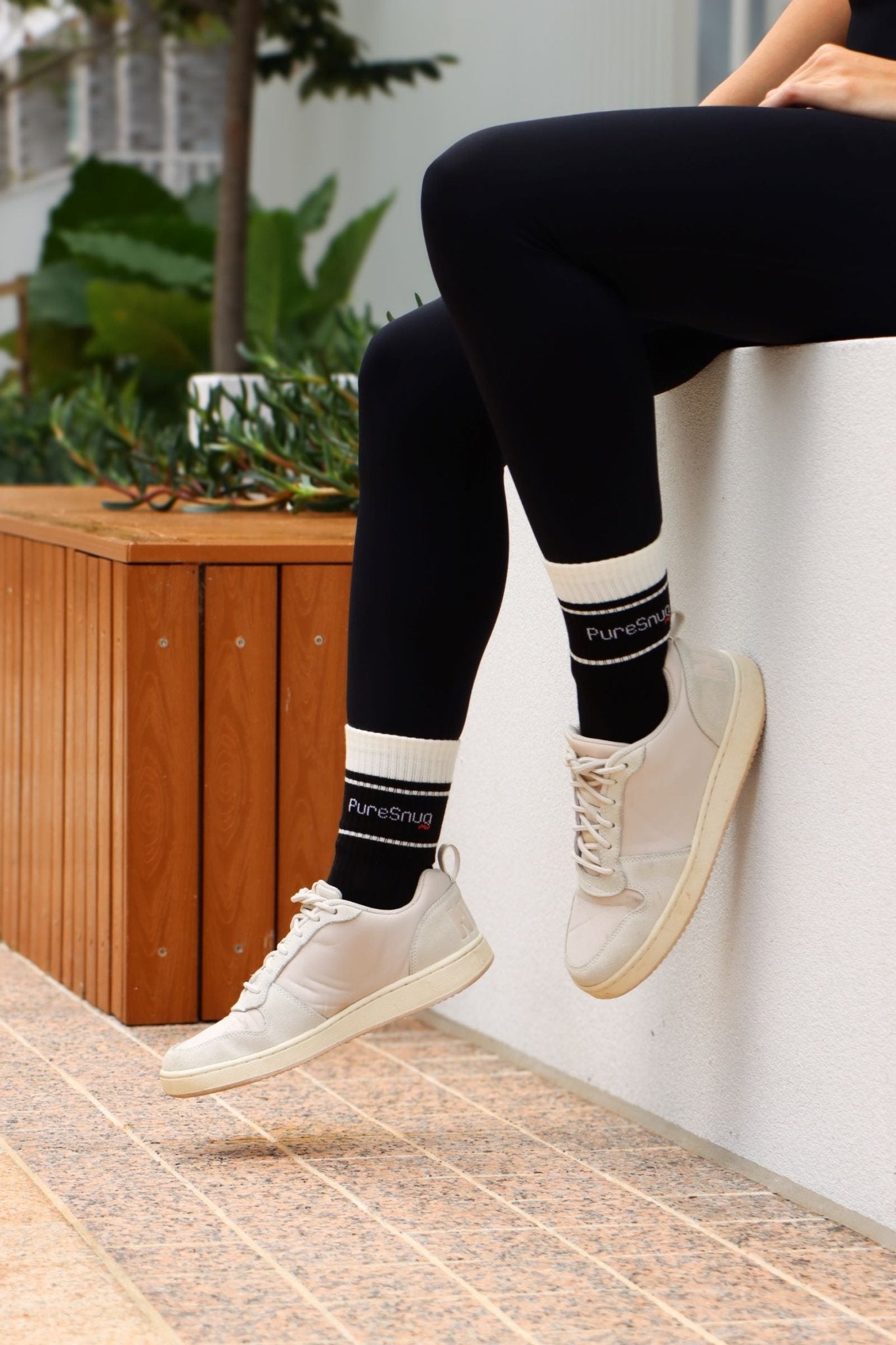A person wearing black leggings and white sneakers sits on a wall, showcasing stylish black and white Puresnug socks.