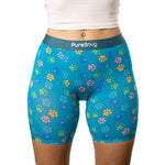 A woman wearing boxer shorts with colorful paw print patterns stands with a white crop top. The tone is playful and vibrant.