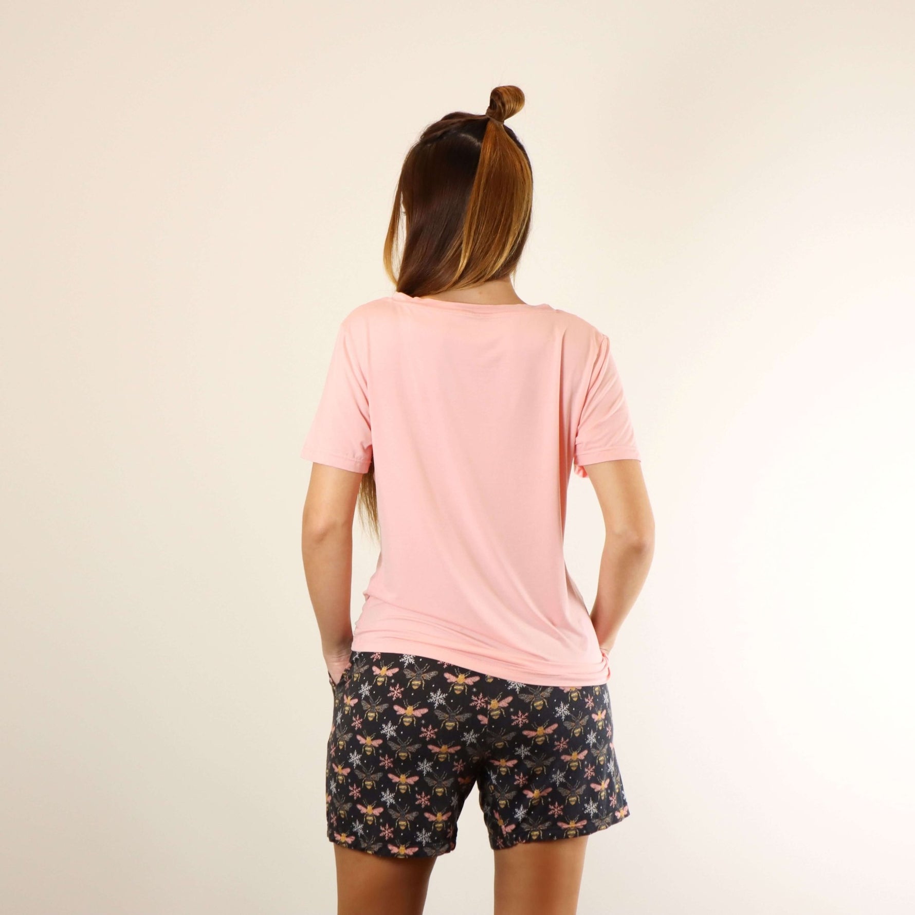 A woman wears a soft v-neck Tee in pink colour, and a soft lounge shorts in bees prints, with hands in pockets, back view.