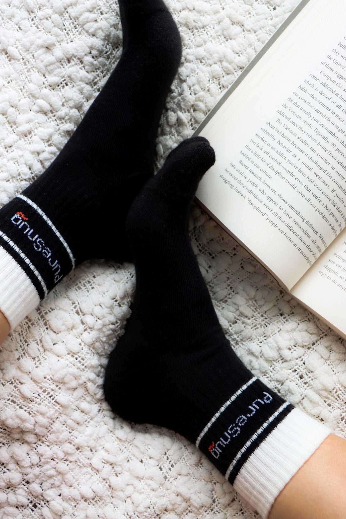 Close-up of black socks with white cuffs and PureSnug branding, resting on a textured blanket beside an open book.